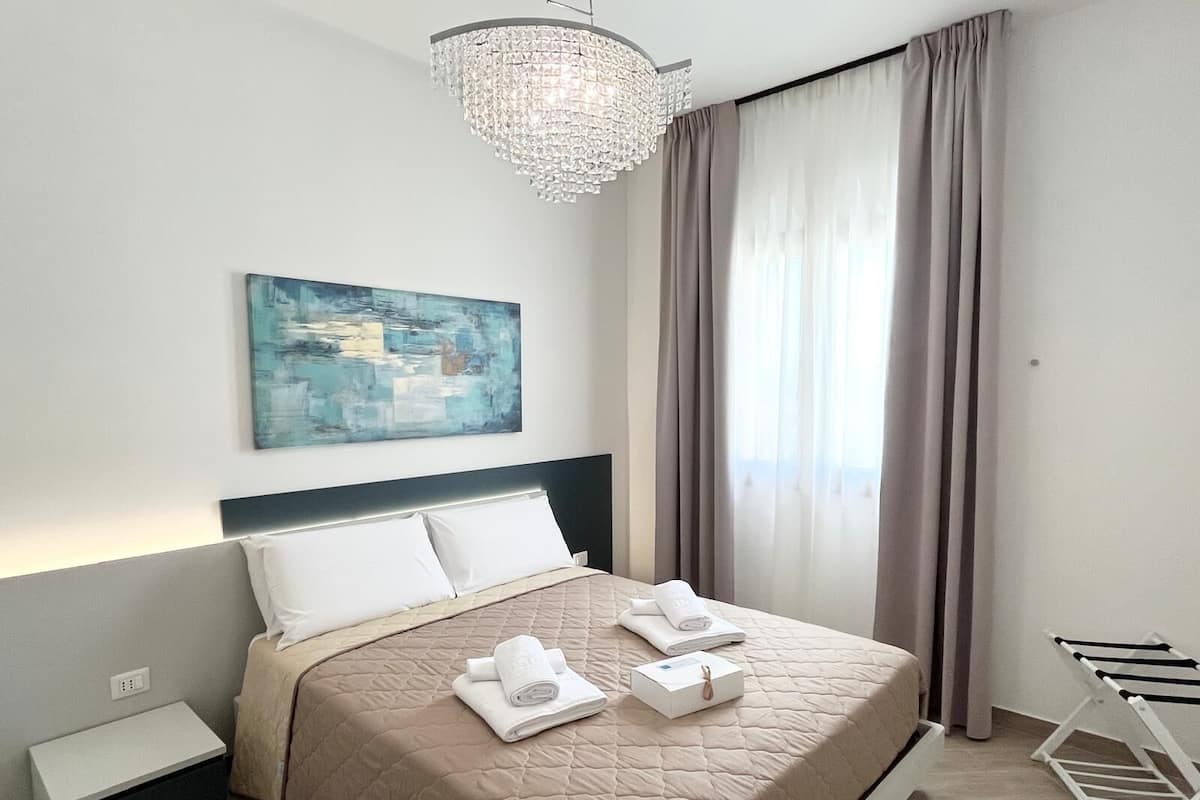 Une chambre d’hôtel moderne avec un grand lit, une table de chevet, un lustre suspendu, un tableau au mur et une fenêtre avec des rideaux.