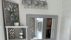Un bagno con un mobile lavabo bianco, uno specchio, uno scaffale con asciugamani, un cestino e un dispenser per carta igienica.