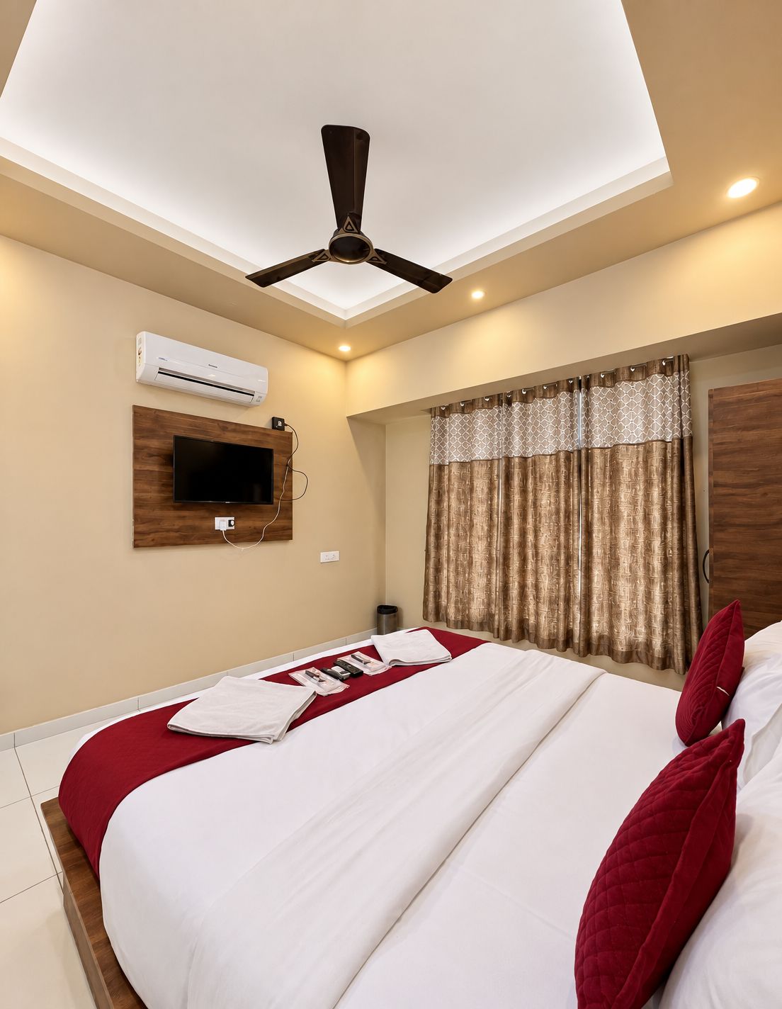 A hotel room with a bed, an air conditioning unit, a ceiling fan, a television, and a window with curtains.