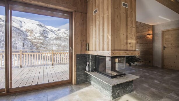 Ein Holzinnenraum mit Kamin aus Stein, einem Balkon mit Blick auf schneebedeckte Berge und einer Holztür.