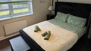 A bedroom with a bed, a window with blinds, a radiator, a bedside table with a lamp, and a wall-mounted mirror. - EV Charger, Airport Access, City Centre (Speke)