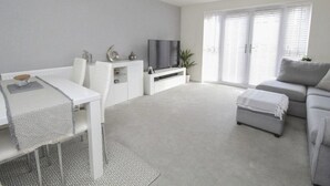 A modern living room with a white sofa, a dining table with a patterned tablecloth, a TV on a white stand, and a large window with blinds. - EV Charger, Airport Access, City Centre (Speke)