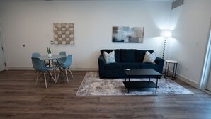 A living room with a dark blue sofa, a coffee table, a dining table with chairs, and a small round table with a lamp. - The Beehive Loft Gym Parking Garage (Salt Lake City)