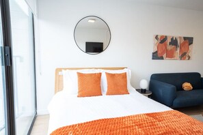 A modern hotel room with a large bed, orange throw pillows, a round mirror, and a blue sofa.