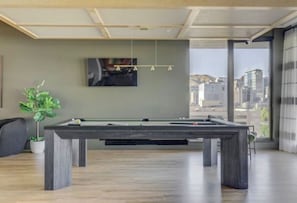 Une table de billard dans une pièce dotée d’une grande fenêtre, d’une plante en pot et d’un téléviseur fixé au mur.