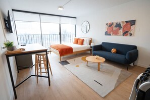 Eine moderne Wohnung mit einem Bett, einem blauen Sofa, einem Essbereich mit Tisch und Stühlen, einem kleinen runden Tisch und einem großen Fenster mit Blick.