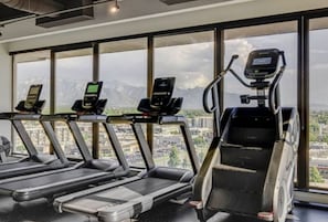 Une salle de sport équipée de tapis de course et d’un appareil de marche en escalier, avec de grandes fenêtres offrant une vue sur la ville.