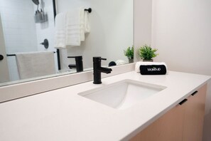 Ein modernes Badezimmer mit einem weißen Waschbecken, einem schwarzen Wasserhahn und einem Spiegel, der ein Handtuch und eine Pflanze reflektiert.