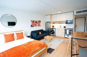 Ein kompaktes Hotelzimmer mit Bett, Sofa, einer kleinen Küchenzeile inklusive Mikrowelle, Herd und Waschmaschine sowie einem runden Spiegel an der Wand.