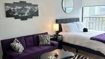 Quarto de hotel moderno com cama, sofá roxo, uma mesinha e um tapete listrado em preto e branco.