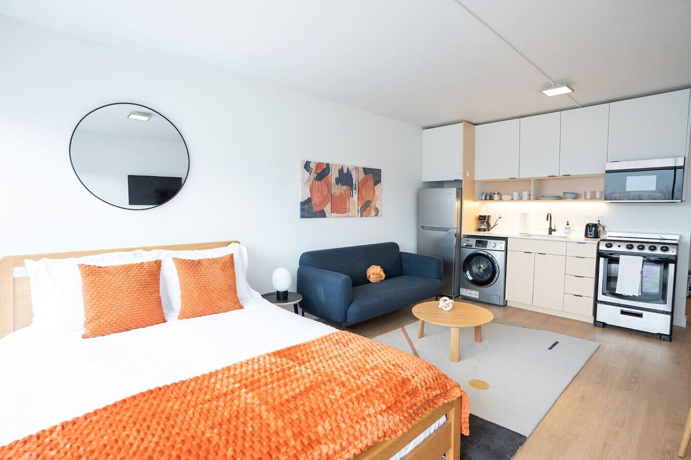 Een moderne hotelkamer met een bed, een sofa, een salontafel, een kitchenette met apparatuur, en een ronde spiegel aan de muur.