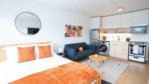 Una habitación de hotel moderna con una cama, un sofá, una mesa de centro, una zona de cocina con electrodomésticos y un espejo redondo en la pared.