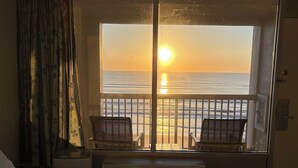 A room with a bed, a window with a view of the ocean at sunset, a radiator, and two chairs by the window. - Cozy studio condo with direct ocean view in amazing Daytona Beach (Daytona Beach)