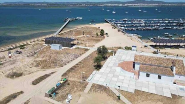 Oversigtsbillede af et kystområde med en lysthavn, sandstrand og en stor bygning med solpaneler.