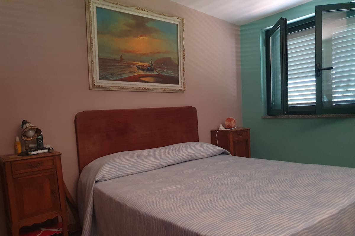 A bedroom with a bed, a nightstand, a painting, and a window with blinds.