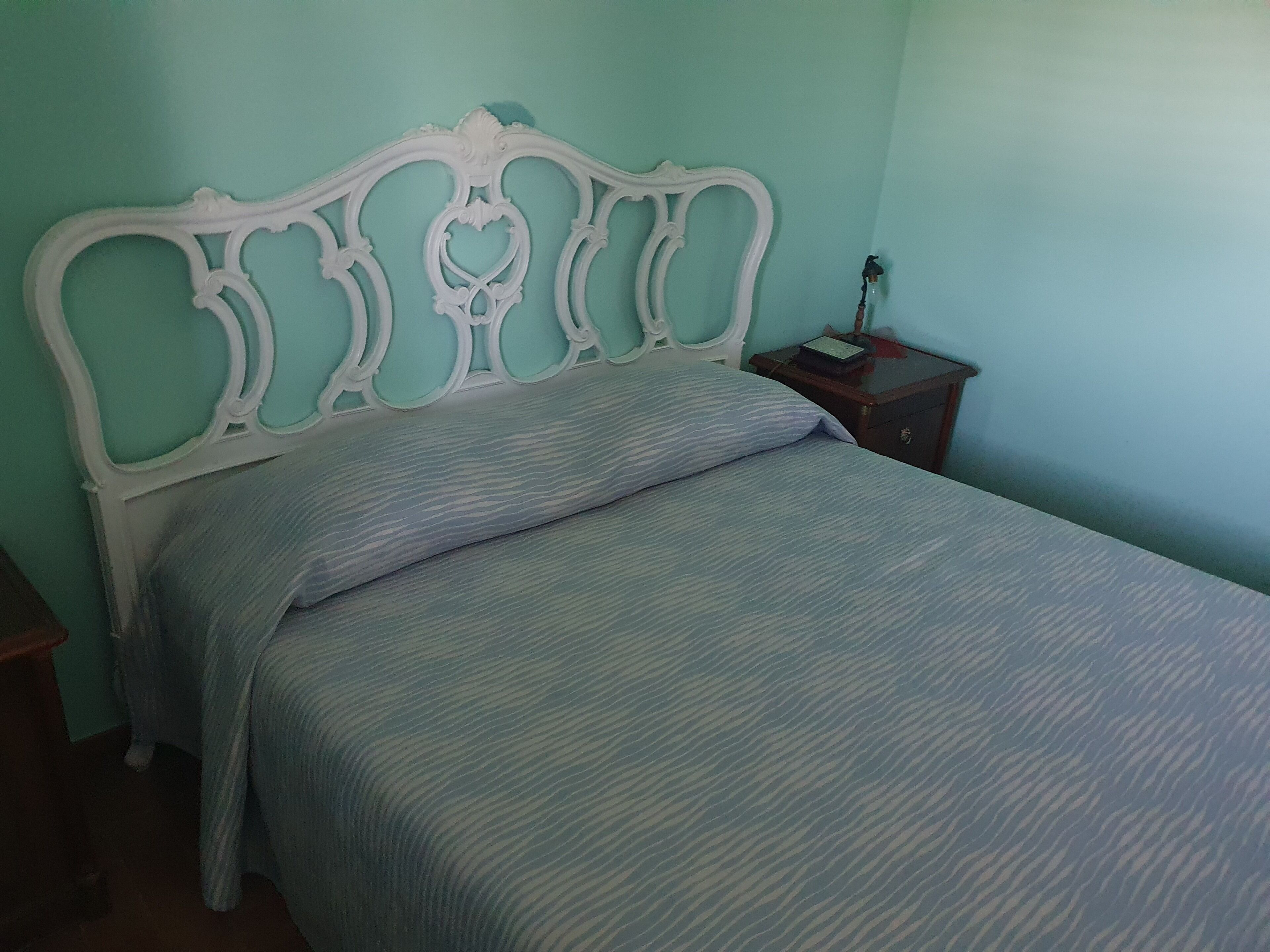 Ein Bett mit weißem Kopfteil, ein Nachttisch mit Lampe und eine Wand mit strukturierter Oberfläche.