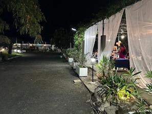 Un espacio para eventos al aire libre con una zona de estar cubierta, plantas en macetas y un camino que lleva a un edificio.