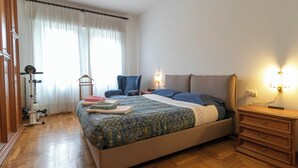Un dormitorio con cama, mesitas de noche, una silla, una máquina de ejercicios y una ventana con cortinas.