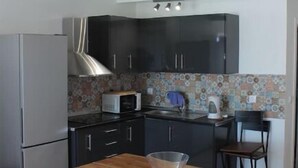 Een kleine keuken met donkere kastjes, een magnetron en een koelkast. Er staat een houten tafel met een geweven bankje.