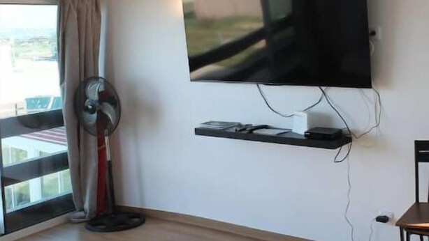 Een moderne woonkamer met een flatscreen televisie aan de muur, een staande ventilator, een houten salontafel en een sofa met een patroon.