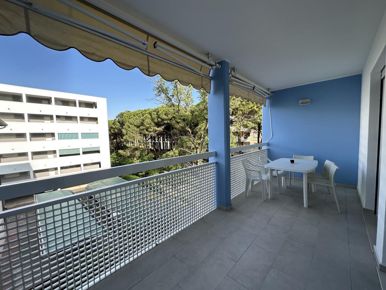 Ein Balkon mit weißem Tisch und Stühlen, einem Metallgeländer und Blick auf ein Gebäude und Bäume.