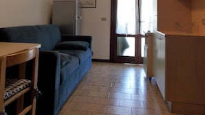 A living room with a sofa, a wooden table, a ceiling fan, and a kitchen area with cabinets and a refrigerator. - Residence Athena-two room apartment 26 B4*++ for 4 persons (Bibione)