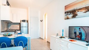 Una cocina moderna con armarios blancos, microondas y refrigerador. Hay un comedor con sillas azules y una mesa. Un televisor se encuentra a la derecha, mostrando un evento de ciclismo.