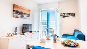 Una habitación moderna con balcón, comedor con mesa lista para dos personas y un televisor a la izquierda.