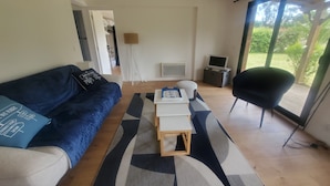 A modern living room with a blue sofa, a small wooden table, and a black chair. There is a large window with a view of greenery outside. - Chalet Lesconil (Plobannalec-Lesconil)