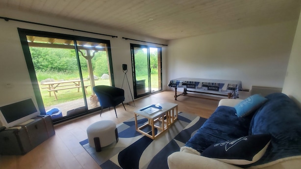 A spacious living room with a large sofa, a coffee table, and a TV. There is a sliding glass door leading to an outdoor area with a table and chairs. - Chalet Lesconil (Plobannalec-Lesconil)