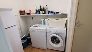 A laundry room with a washing machine, dryer, and a shelf with various items. - Chalet Lesconil (Plobannalec-Lesconil)