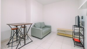 A small, sparsely furnished room with a grey sofa, a small table with stools, a TV on a stand, and a shelf unit.