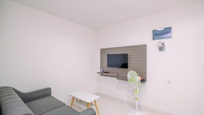 A modern living room with a grey sofa, a small white table, a mounted TV, a standing fan, and a wooden shelf. - Comfort and Homey 2BR at Sentul Tower Apartment (Citaringgul)