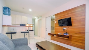 A modern living room with a sofa, a wooden coffee table, a TV mounted on a wooden panel wall, and a kitchen area in the background. - Spacious 2BR at Sentul Tower Apartment (Citaringgul)