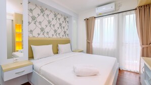 Una cama bien hecha con ropa de cama blanca y una toalla doblada, una mesita de noche con una lámpara, un aire acondicionado instalado en la pared y una ventana con cortinas traslúcidas.