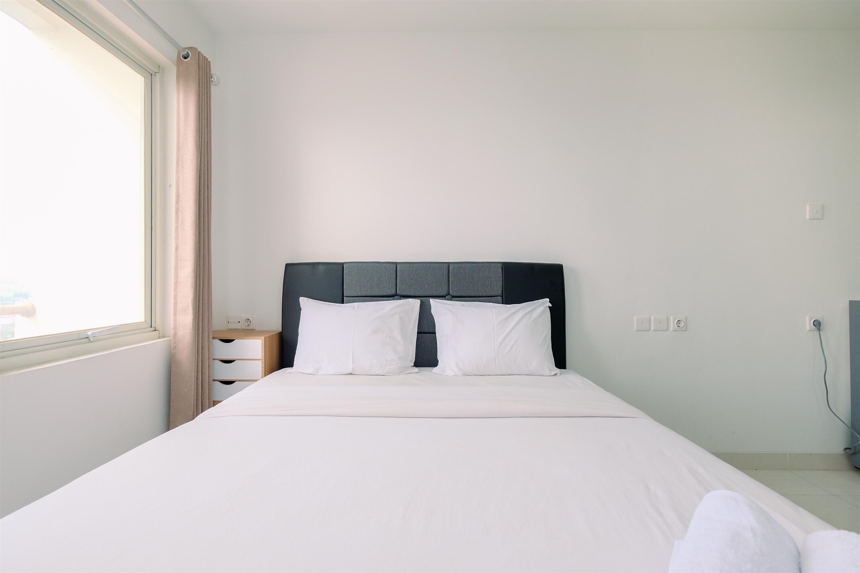 A neatly made bed with white linens and two pillows, a bedside table with a lamp, and a window with curtains.