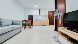 Una sala de estar con un sofá, un televisor y un ventilador. Se observa una cocina al fondo con armarios y un refrigerador.