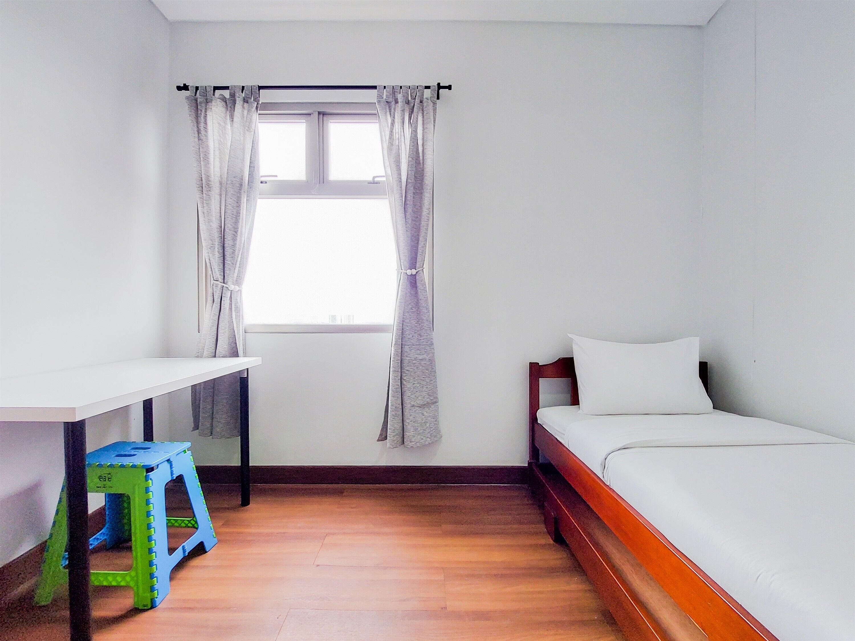 Eine kleine, einfache Unterkunft mit Holzboden, einem weißen Bett, einem kleinen Schreibtisch und einem Fenster mit Vorhängen.