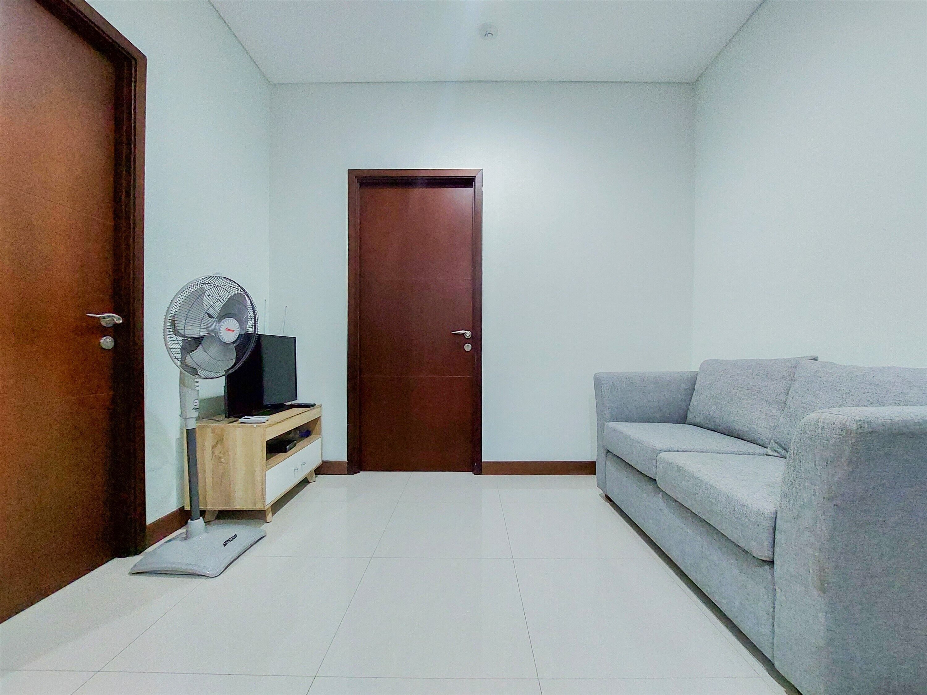 A living room with a grey sofa, a wooden cabinet with a TV and fan, a closed wooden door, and a tiled floor.