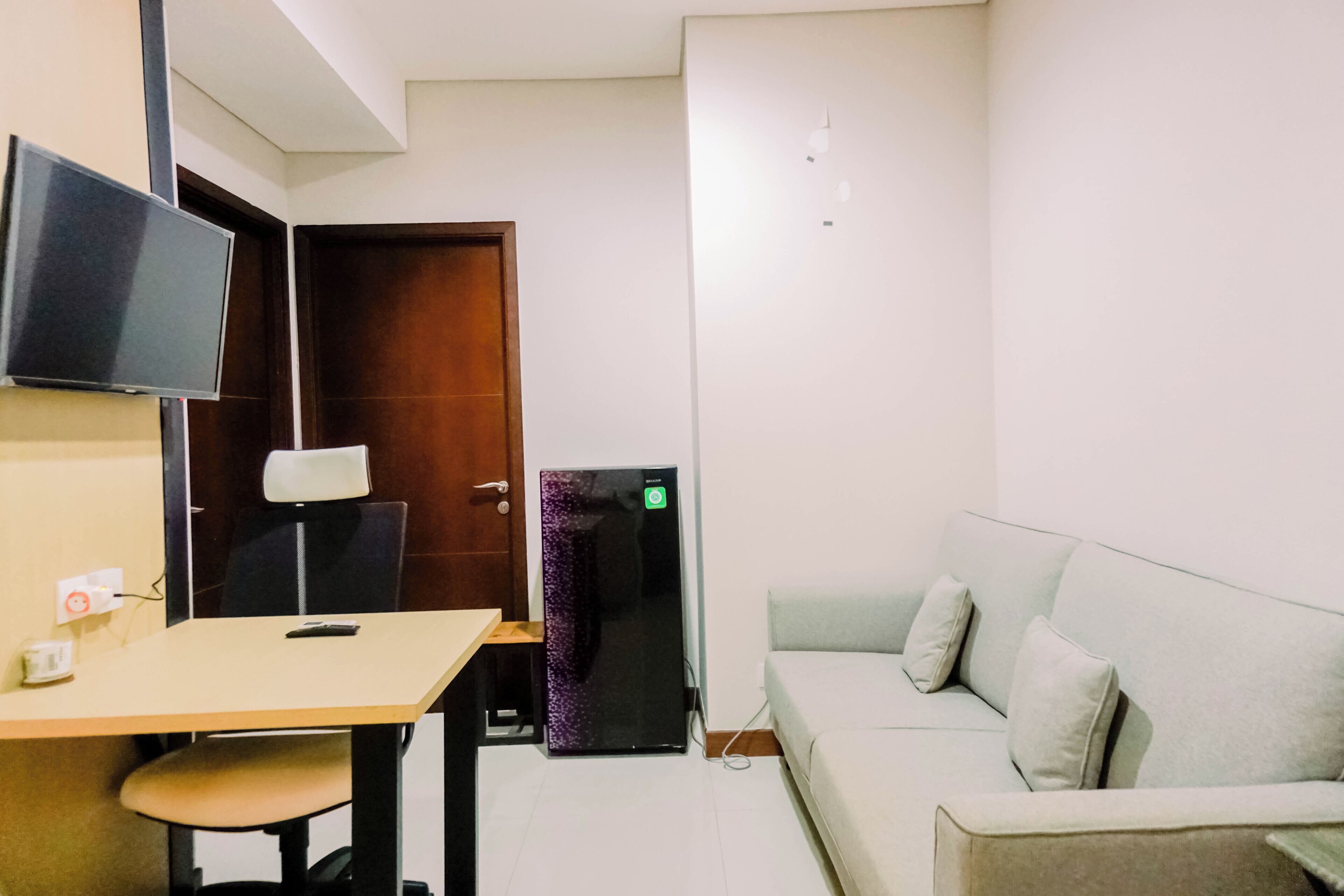 Una habitación pequeña y moderna con una puerta de madera, un televisor de pantalla plana montado en la pared, una mesita con una silla y un sofá de color claro.