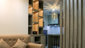 A modern living room with a sofa, wooden shelving, and a kitchen area in the background. - Fancy and Nice 2BR at Sentul Tower Apartment (Citaringgul)