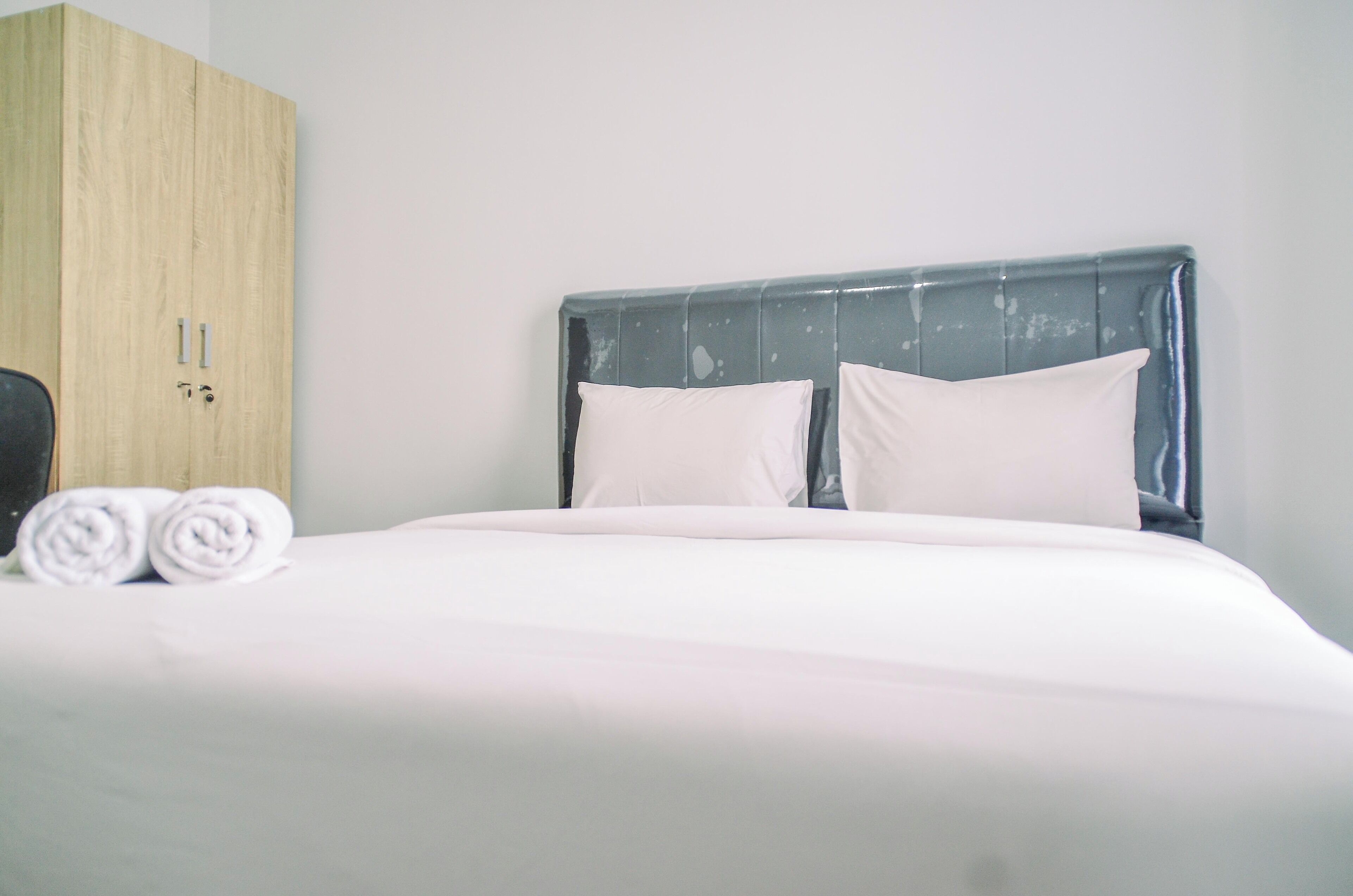 Una cama bien hecha con sábanas y almohadas blancas, un armario de madera con dos pomos y una pared lisa.