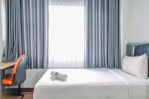 Una habitación de hotel con cama, escritorio, una silla naranja y ventana con cortinas.
