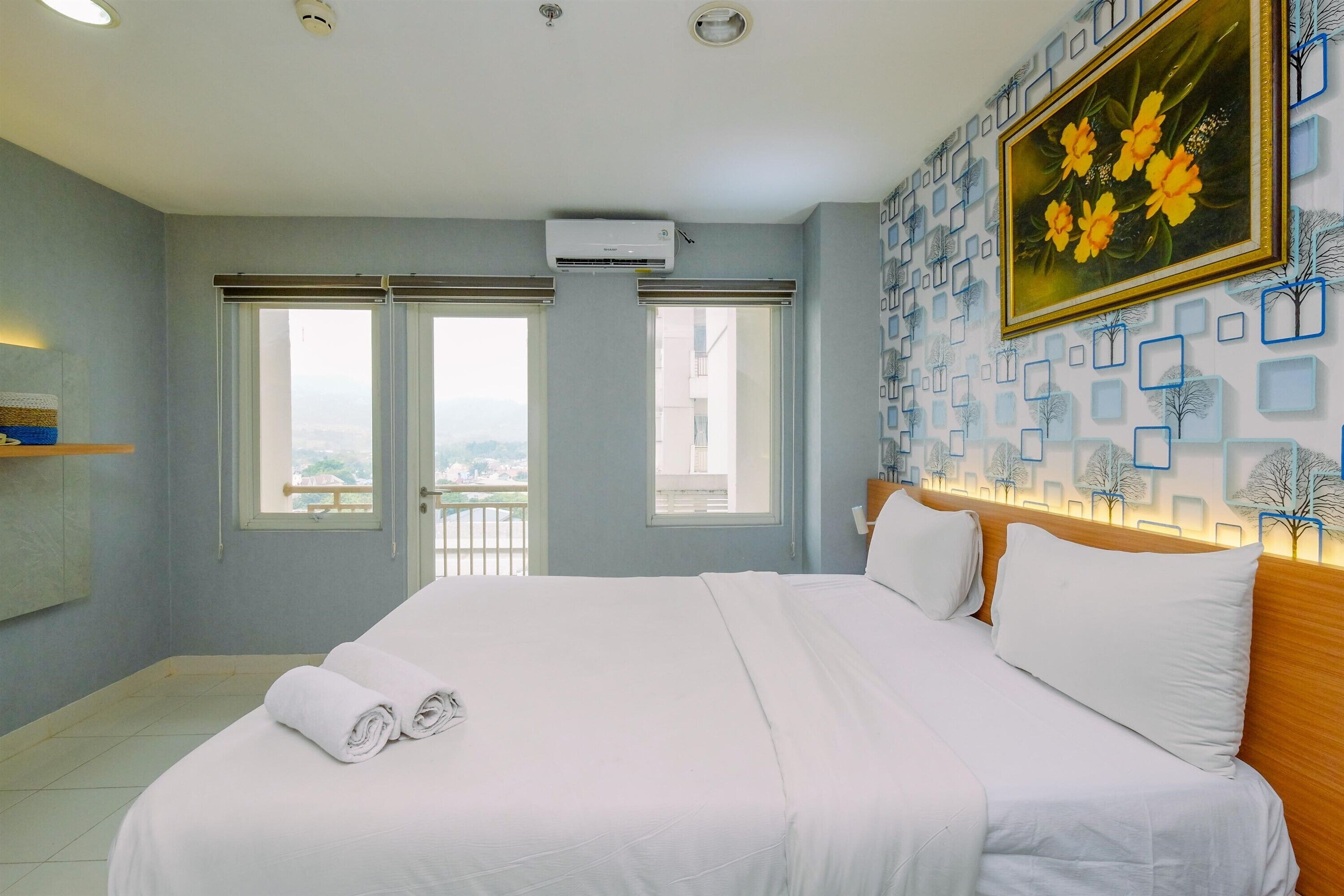 A hotel room with a large bed, two windows, an air conditioning unit, and a wall with a floral painting.