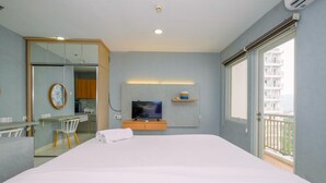 A hotel room with a large bed, a TV on a wooden shelf, a small table with a chair, and a balcony with a view of buildings. - Cozy Studio at Sentul Tower Apartment (Citaringgul)