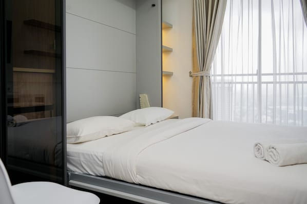 Une chambre d’hôtel moderne avec un grand lit, une cloison en verre et une vue sur la ville à travers de fines rideaux.
