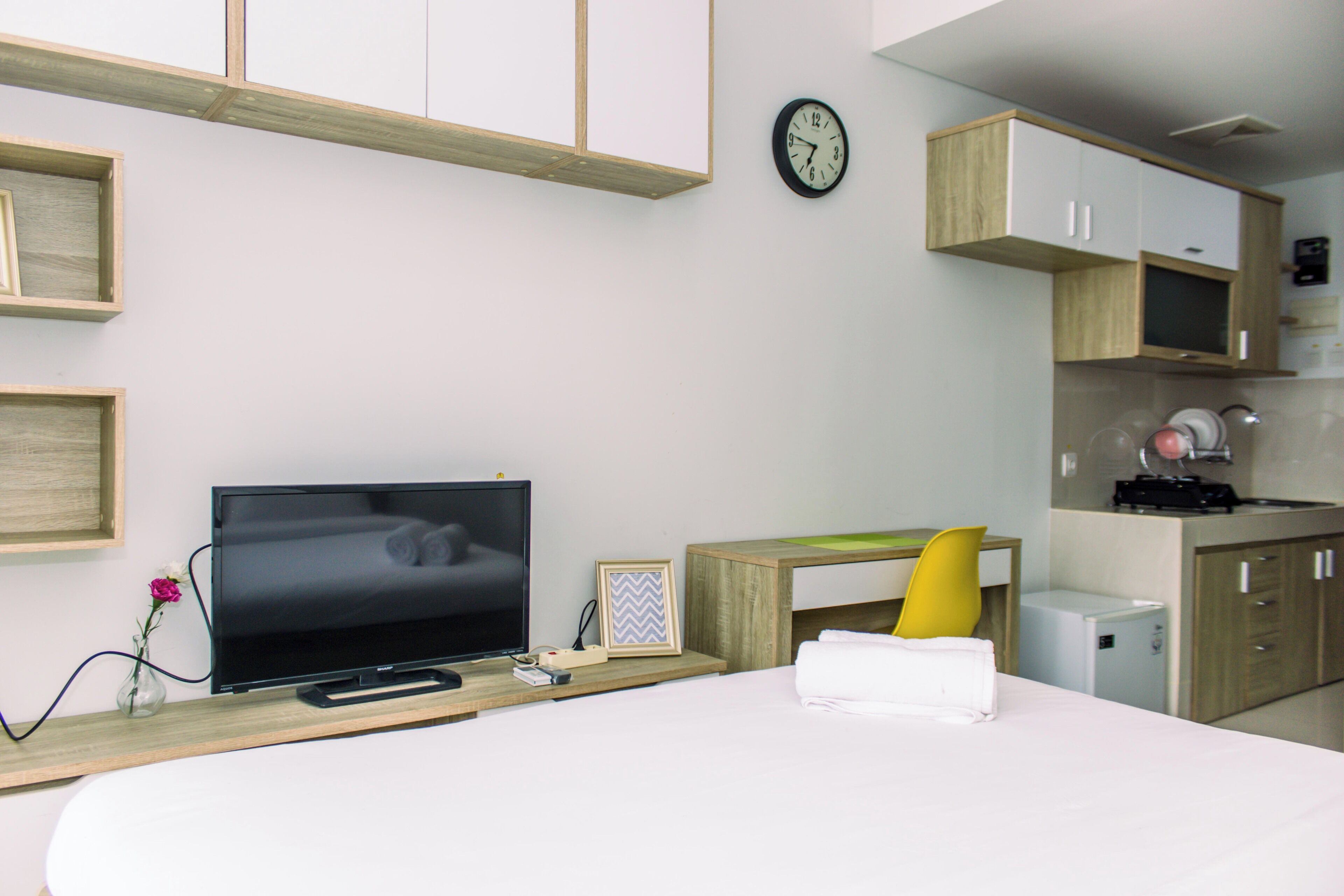Ein modernes Hotelzimmer mit Bett, Fernseher, Schreibtisch mit Computer, einer kleinen Küchenzeile und einer Wanduhr.