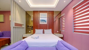 A modern hotel room with a large bed, a purple sofa, a wooden wardrobe, and a window with red blinds. - Homey and Modern Studio at Sentul Tower Apartment (Citaringgul)