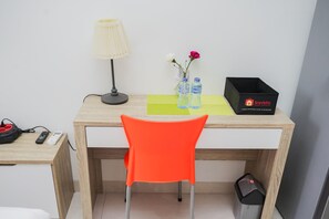 Ein Hotelzimmer mit einem Holztisch, einem orangefarbenen Sessel, einer Lampe, einem Mülleimer und einer Reisetasche.