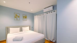 A neatly made bed with white linens and a rolled towel, a grey headboard, and a wall-mounted air conditioner. - Modern Look 2BR at Sentul Tower Apartment (Citaringgul)